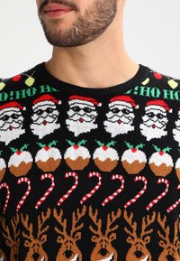 Homme portant un pull de Noël avec des motifs de visages du Père Noël, pudding, cannes de bonbon et rennes en rouge, blanc, brun et vert sur fond noir.