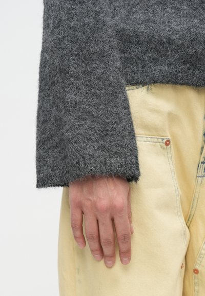 Pulover din lână gri cu manșetă largă, purtat peste pantaloni din denim galben deschis. Țesătura texturată are un aspect moale și pufoasă.