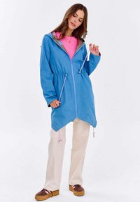 Kobieta poprawiająca sznurki w niebieskiej kurtce z kapturem zapinanej na zamek, noszonej na różowej koszuli, beżowych spodniach i brązowych sneakersach na platformie.