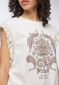 T-shirt bianca in cotone con maniche frangianti. Presenta una stampa floreale e paisley marrone con il testo "SEGUILO CON IL FLUSSO" sotto il design.