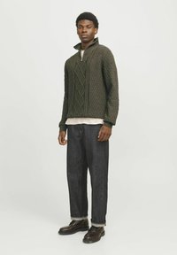 Žalias megztinis su diamantino raštu, su užtrauktuku ant apykaklės; derinamas su tamsiai mėlynomis džinsomis ir juodais laiduojamais batais. Modelis stovi prieš paprastą foną.