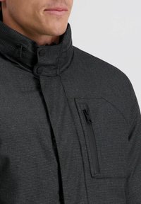 Graue mit Reißverschluss versehene Jacke mit hohem Kragen, strukturiertem Stoff und schwarzen Akzenten, einschließlich einer Brusttasche mit Reißverschluss.