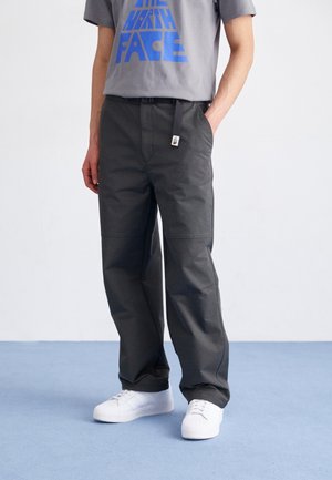 Persoon draagt donkergrijze broek met een zwarte riem en witte sneakers, staand op een lichtblauwe vloer tegen een effen witte muur.