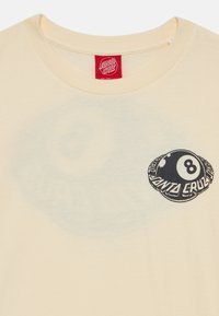 Kremkrāsas T-kreklis no kokvilnas, ar melnu astotā bumbiņas grafiku uz krūšu daļas. Apaļš kakla izgriezums un īsās piedurknes.