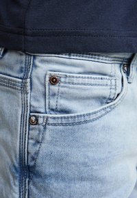 Jeans din denim bleu deschis, cu buzunar aplicat, nasturi de cupru și cusături vizibile. Alături, un top din material închis la culoare.