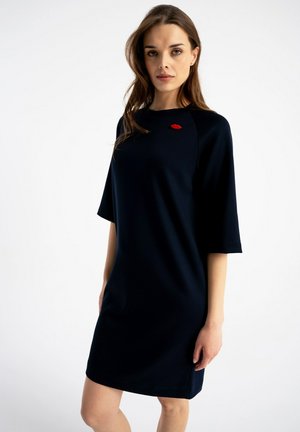 Kobieta ubrana w prostą czarną sukienkę do kolan z rękawami do łokcia i małym czerwonym wzorem ust w pobliżu dekoltu.