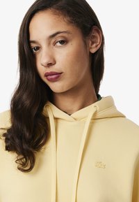 Gelbe Kapuzenjacke mit weichem Material, ausgestattet mit einer Kordelzugkapuze und einem kleinen gestickten Logo. Entworfen mit einem lockeren Schnitt und einer Vordertasche.