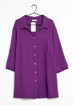 Lilla tekstureret bluse med lange ærmer, krave og seks knapper foran, hængende på en sort bøjle mod en hvid baggrund.