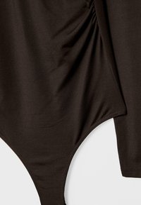 Detailní záběr tmavě hnědého látkového body s nařaseným švem a zapínáním na patentky na spodním okraji.