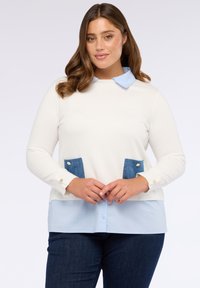 Femeie cu păr ondulat brunet, îmbrăcată cu un pulover alb cu buzunare albastre peste o cămașă cu guler albastru deschis și blugi închiși, stând cu mâinile împreunate.