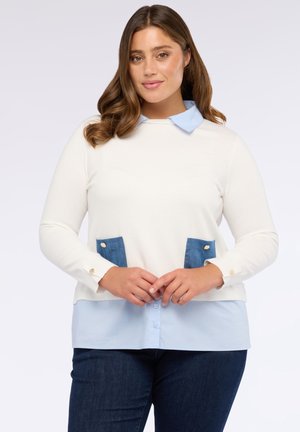 Donna con i capelli castani mossi che indossa un maglione bianco con tasche blu su una camicia collare blu chiaro e jeans scuri, in piedi con le mani giunte.