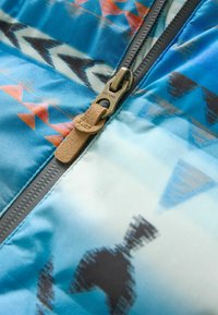 Sección de chaqueta azul con patrones que presentan intrincados diseños geométricos en naranja y negro. Un zipper de bronce con una tira de cuero color beige es visible.