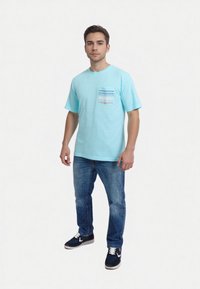 Camiseta a rayas azul claro con un bolsillo multicolor, combinada con jeans azules y zapatillas oscuras. La tela parece suave y casual.