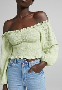 Blusa verde chiaro, con spalle scoperte, in tessuto texturizzato e bordi arricciati, abbinata a jeans in denim blu a vita alta.