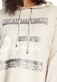 Beige hoodie med silverglitter och vit text som läser "SKAPA LYCKA," "SPRIDA," "GLAD LIV," "Optimism," och "INSPIERA."