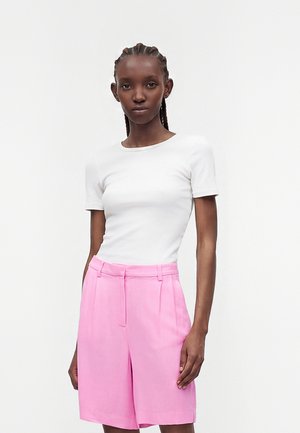 Femme aux cheveux tressés portant un haut blanc à manches courtes et un short rose ample taille haute, debout devant un fond uni.