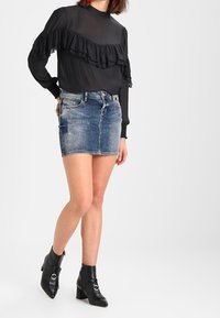 Femme portant un blouse noire à manches longues avec volants, une jupe en denim bleu courte et des bottines noires avec des boucles argentées, se tenant contre un fond uni.