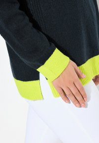 Mörkstickad tröja med ljusgula ribbade manschetter och nederkant. Har sidofickor och en mjuk textur. Bärs med vita byxor.