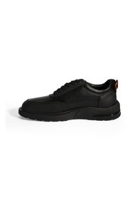 Sneakers en cuir noir avec une tige texturée, bout rond, design à lacets et une semelle noire épaisse avec un petit onglet de talon orange.