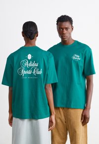 Teal bomulds t-shirt med "Adidas Sports Club" tryk i hvid på fronten og bagsiden, med en løs pasform og rund halsudskæring.