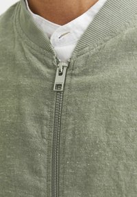 Grüne Bomberjacke mit Reißverschluss, geripptem Kragen und strukturiertem Stoff. Ein weißes Hemd ist unter der Jacke sichtbar.