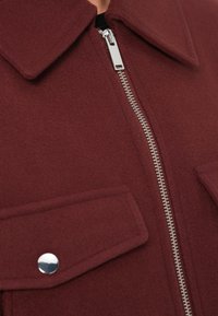 Μπορντό μάλλινο σακάκι με ασημί φερμουάρ, μεγάλο γιακά και δύο τσέπες στο στήθος με κουμπιά snap. Η υφή της επιφάνειας ενισχύει τη ζεστασιά.