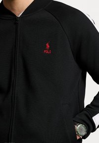 Černá bunda na zip s žebrovaným límcem, s červeně vyšívaným logem "Polo" a bílými akcenty na rukávech. Vyrobená z texturované látky.