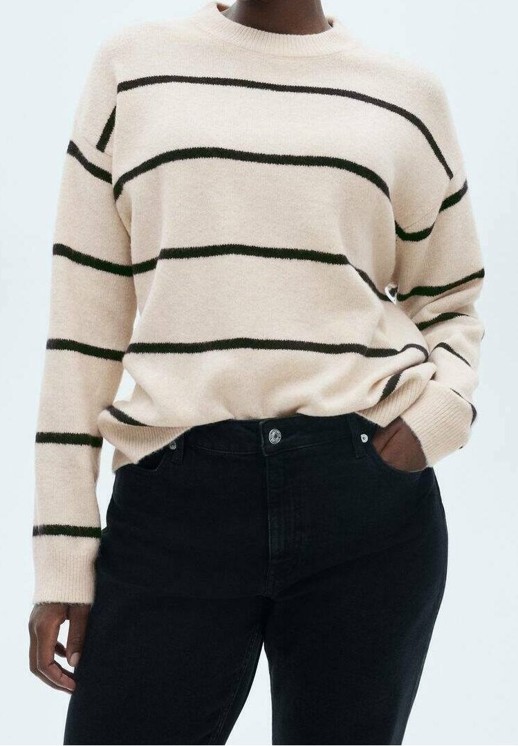 Personne portant un pull beige avec des rayures horizontales noires et un pantalon noir, debout avec une main dans la poche devant un fond uni.