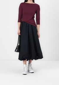 Burgundy topp med trekvartsärmar, i kombination med en svart, voluminös midikjol och vita plattformssneakers, som håller en liten svart väska.