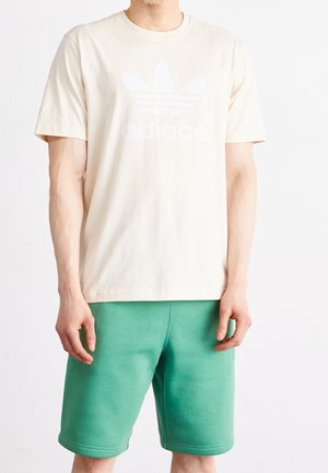 Homme portant un t-shirt Adidas écru avec logo et un short vert, debout devant un fond blanc uni.
