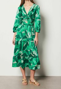 Kvinna klädd i en grön blommig midiklänning med 3/4-ärmar, vriden frontdetalj och gyllene platta sandaler, ståendes mot en vanlig vägg.