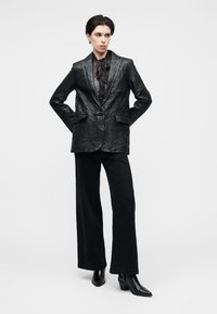 Μαύρο δερμάτινο σακάκι με γιακά με εγκοπές, κλείσιμο με ένα κουμπί και δύο μπροστινές τσέπες. Συνδυασμένο με μαύρο παντελόνι ψηλόμεσο, φαρδύ στις μπατζάκια, και μυτερές μπότες.