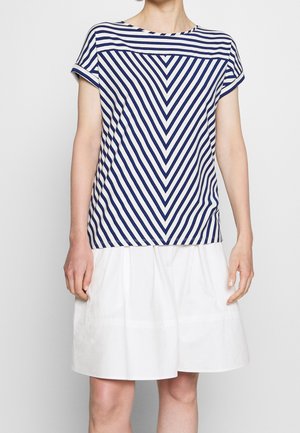 Vrouw in een korteklepen gestreept shirt met een zigzagpatroon en een witte knie-lange rok tegen een effen achtergrond.