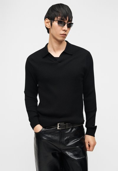 Νεαρός άνδρας φορώντας μαύρο ριμπ μακρυμάνικο πόλο, μαύρο γυαλιστερό δερμάτινο παντελόνι, μαύρη ζώνη και σκούρα ορθογώνια γυαλιά ηλίου μπροστά σε λευκό φόντο.