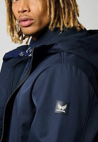 Veste marine avec capuche, dotée d'une fermeture éclair et d'une texture côtelée. Comprend un patch logo Kaporal noir. Bandana bleu noué visible.