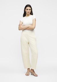 Γυναίκα με μακριά σκούρα μαλλιά, που φοράει λευκό εφαρμοστό T-shirt, κρεμ φαρδιά παντελόνα και μπεζ πλατφόρμες, στέκεται με σταυρωμένα τα χέρια.