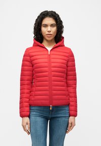 Γυναίκα με σγουρά μαλλιά που φοράει κόκκινο καπιτονέ μπουφάν με φερμουάρ και κουκούλα, και μπλε τζιν, στέκεται μπροστά σε απλό λευκό φόντο.