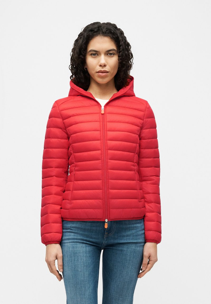 Γυναίκα με σγουρά μαλλιά που φοράει κόκκινο καπιτονέ μπουφάν με φερμουάρ και κουκούλα, και μπλε τζιν, στέκεται μπροστά σε απλό λευκό φόντο.