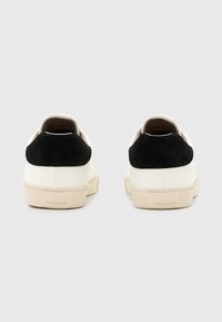 Baskets en cuir blanc avec accents en daim noir au talon, semelle plate et marquage minimal à l'arrière. Texture lisse, design low-top.