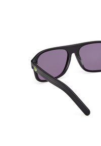 Svarta solglasögon med lila linser, robust plastbåge, avsmalnande skalmar och en grön logodetalj på vänster sida.