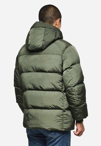 Grüne Puffjacke mit einer Kapuze, die ein gestepptes Design und eine weiche Textur aufweist. Die Rückseite zeigt eine lockere Passform und elastische Bündchen. Getragen mit blauen Jeans.