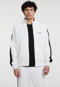 Witte zip-up jas met zwarte zijstrepen, gemaakt van gladde stof. Heeft een hoge kraag en de tekst "GUESS JEANS" op de borst.