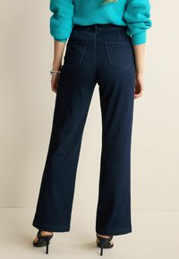 Jean large bleu foncé à taille haute, avec deux poches arrière et des détails de couture subtils. Complété par un pull teal et des talons.