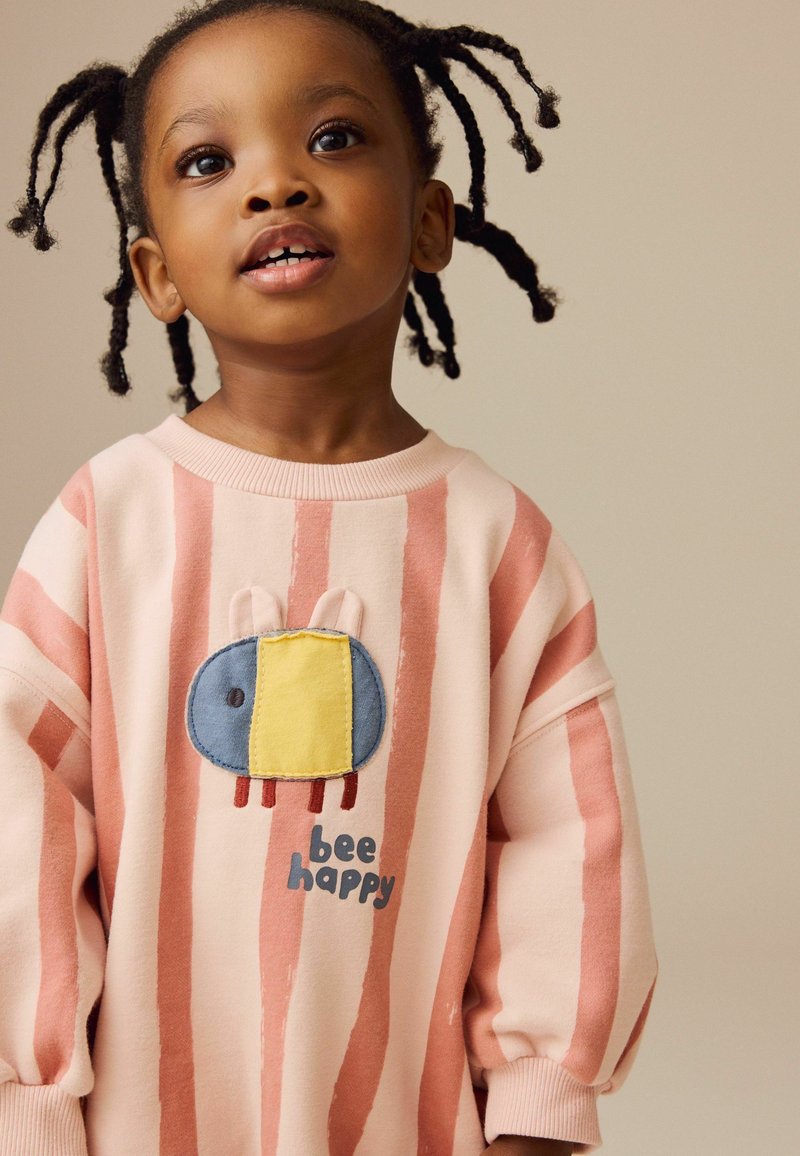 Lyserød sweatshirt med lodrette koralfarvede striber, der har et broderet blå og gul bie-design samt teksten "bee happy" nedenunder.