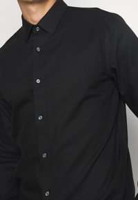 Svart knäppt skjorta med standardkrage, med en slät textur och sex knappar framtill. Enkel design, figurnära skärning.