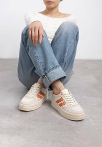 Zapatillas blancas con rayas laterales naranjas, diseño de encaje, suela acolchada y parte superior texturizada. Combinadas con jeans azules claros con dobladillo.