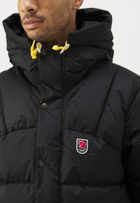 Black puffer jacket with a high collar, featuring yellow drawstrings, large quilted sections, and a logo patch near the chest.
