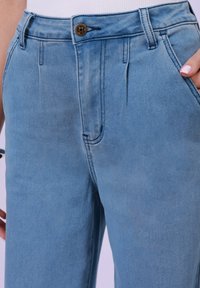 Svijetloplave traperice s visokim strukom, prednjim plavinama i bočnim džepovima. Tkanina ima mekanu teksturu i blago izblijedjeli finiš.