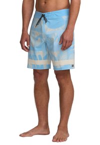 Muž v modrých šortkách s abstraktnými béžovými vzormi a čiernou šnúrkou, stojí bosý na bielom pozadí.