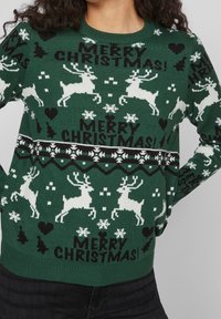 Grüner Strickpullover mit weißen Rentieren und Schneeflocken, akzentuiert mit schwarzem Text "FROHE WEIHNACHTEN!" und gemusterten Rändern.